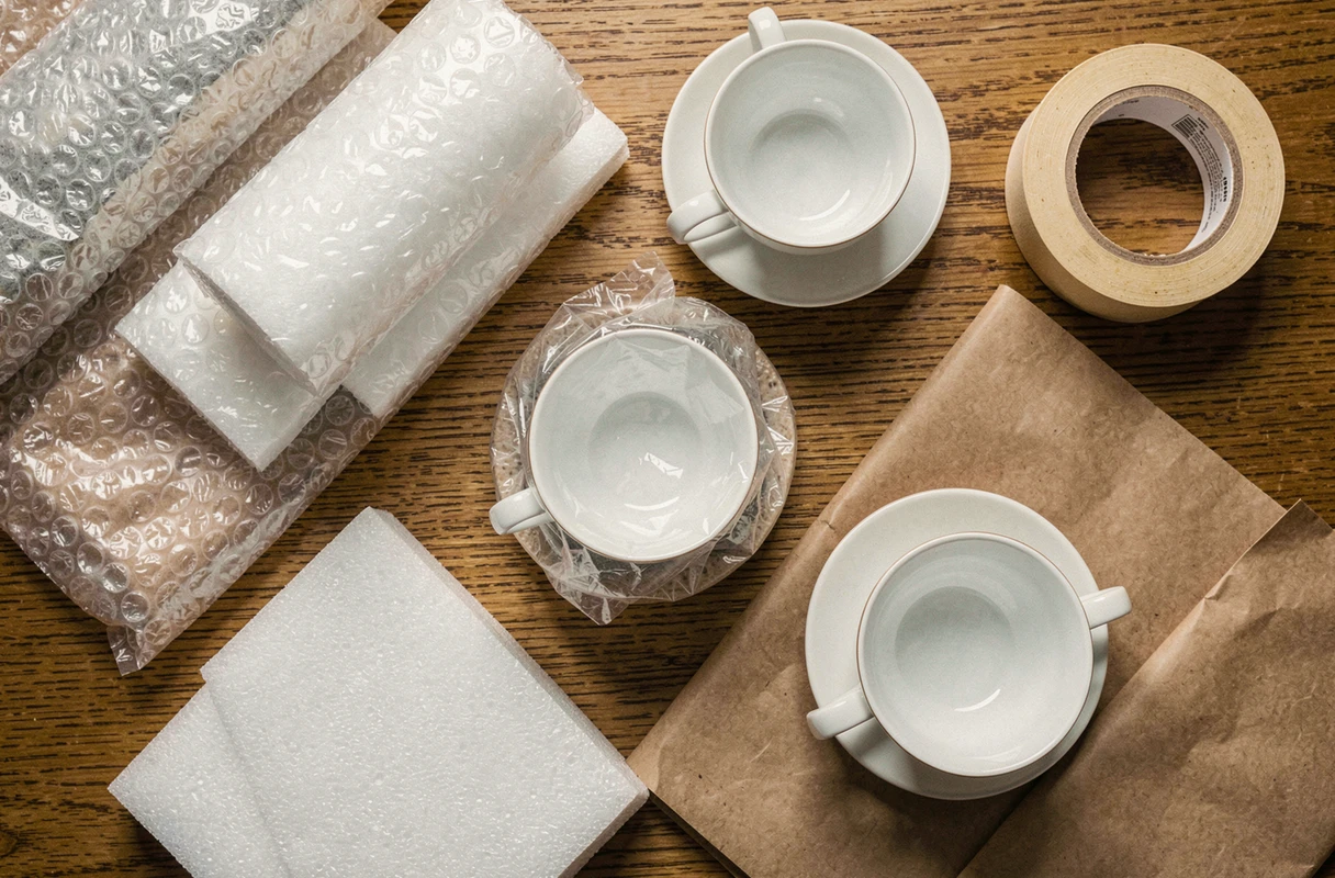 Vue de dessus de matériaux d'emballage pour vaisselle fragile incluant papier bulle, papier kraft et tasses en porcelaine sur une table en bois
